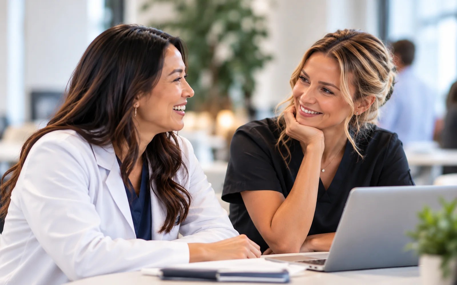 Two smiling dermatology clinicians in scrubs and a white coat talking together at a professional gathering.